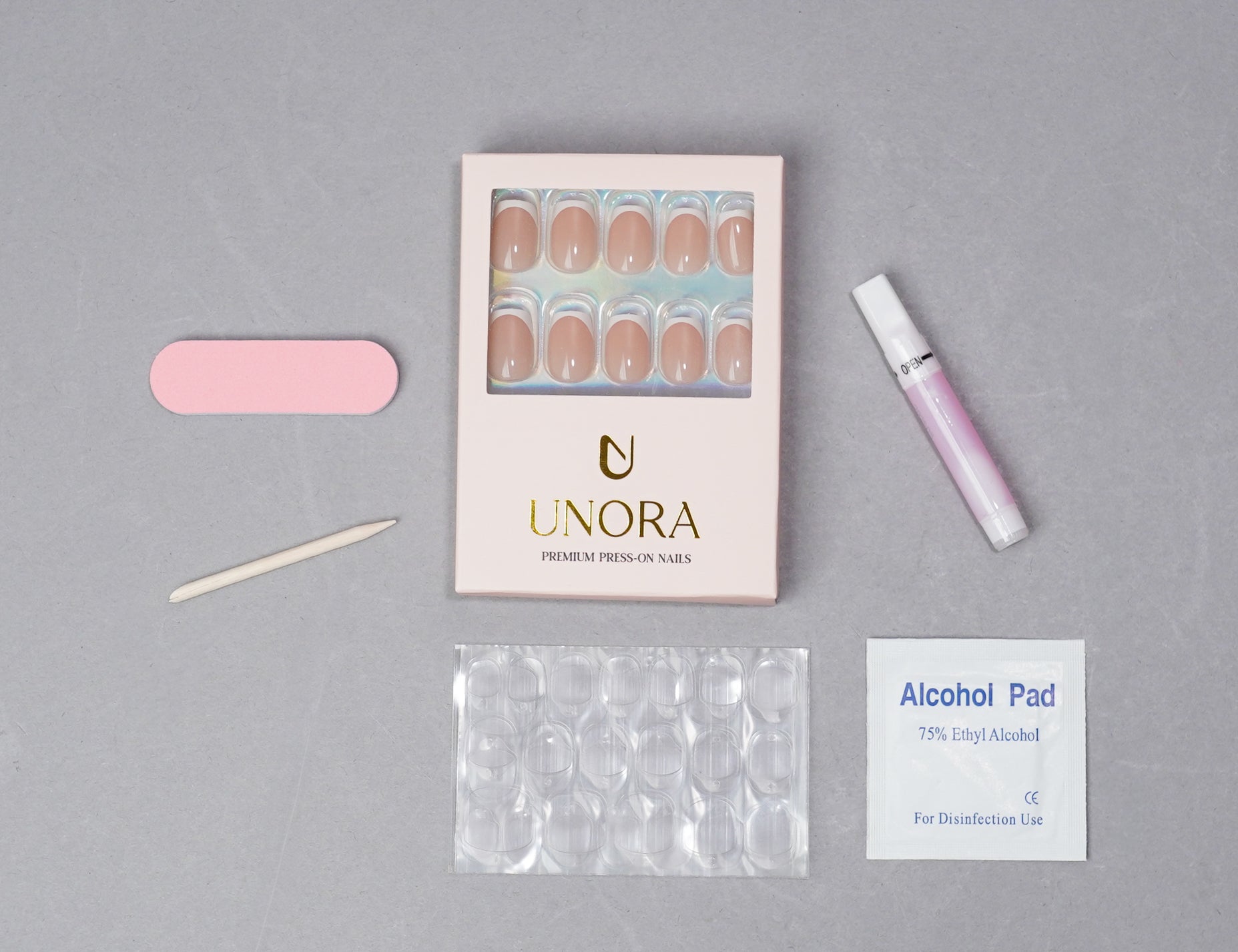 UNORA press-on nail kit on a gray background, including a box of nude false nails, a pink nail file, a wooden nail tool, a pink nail polish tube, a clear nail form pack, and an alcohol pad.