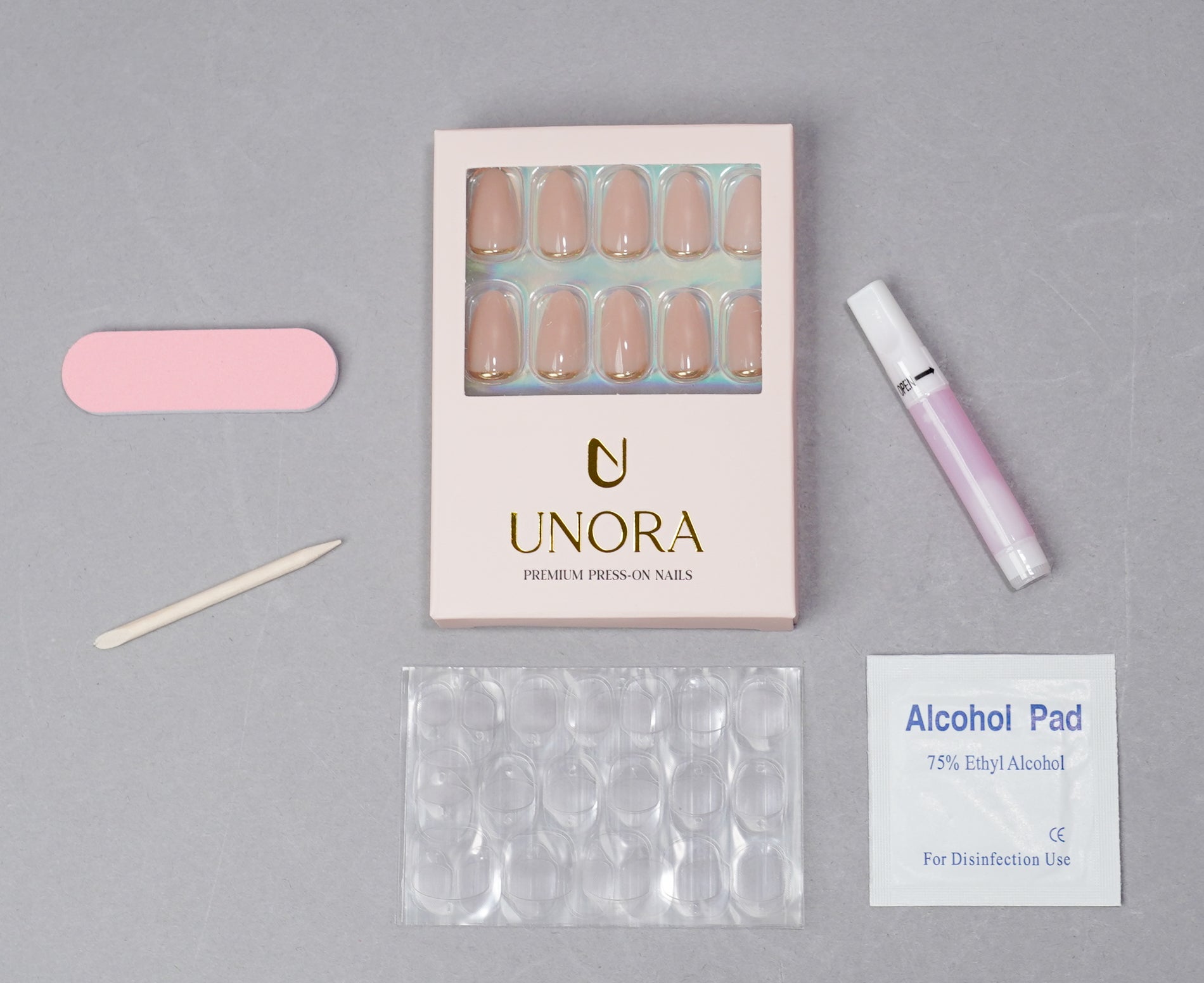 UNORA press-on nail kit on a gray background, including a box of nude beige press-on nails, a pink nail file, a wooden nail tool, a pink nail polish tube, a clear plastic nail form tray, and an alcohol pad.