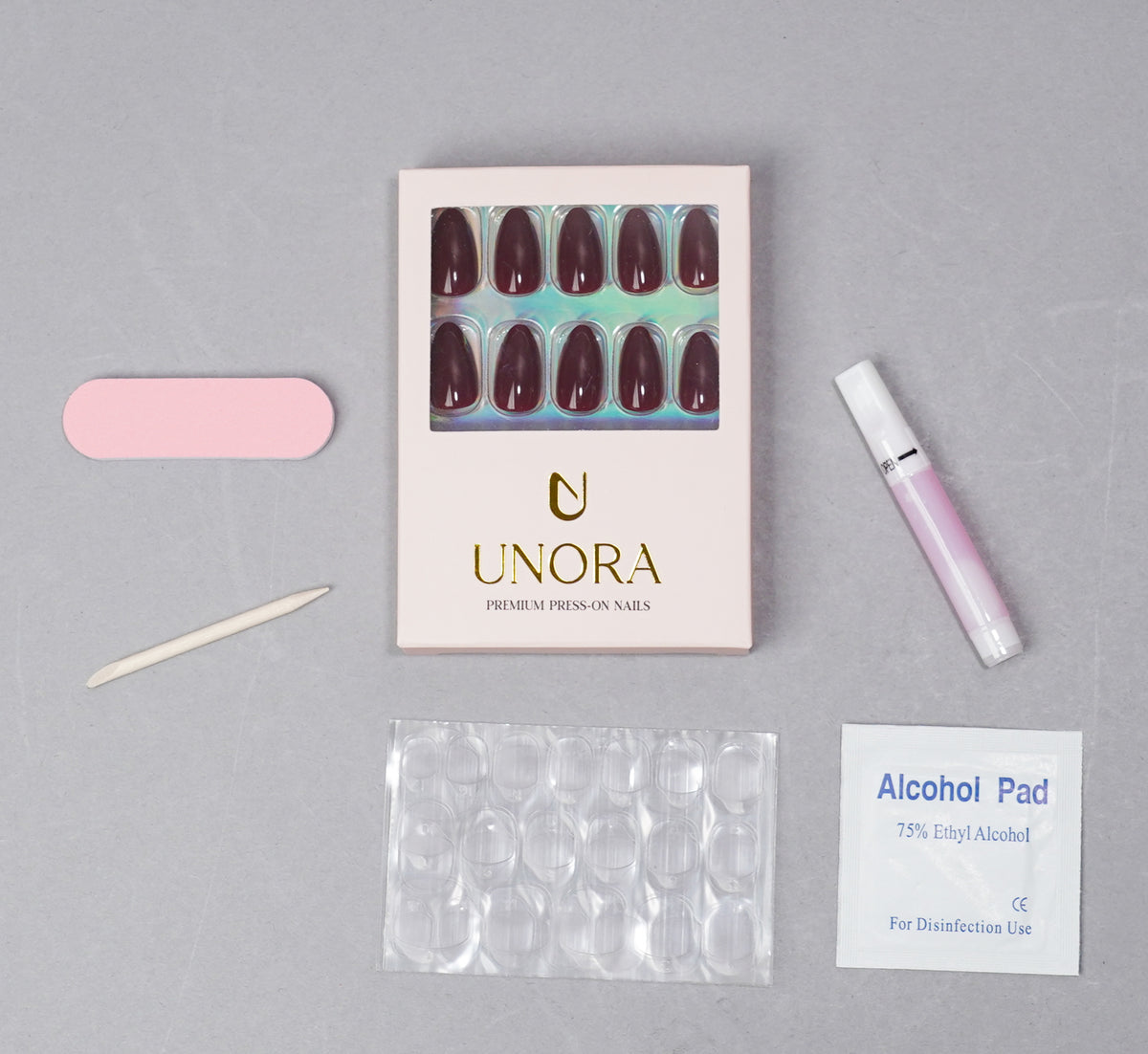 UNORA press-on nail kit arranged on a light gray background, including a box of dark burgundy press-on nails, a pink nail file, a wooden nail tool, a pink nail polish bottle, a clear nail tray, and an alcohol pad.
