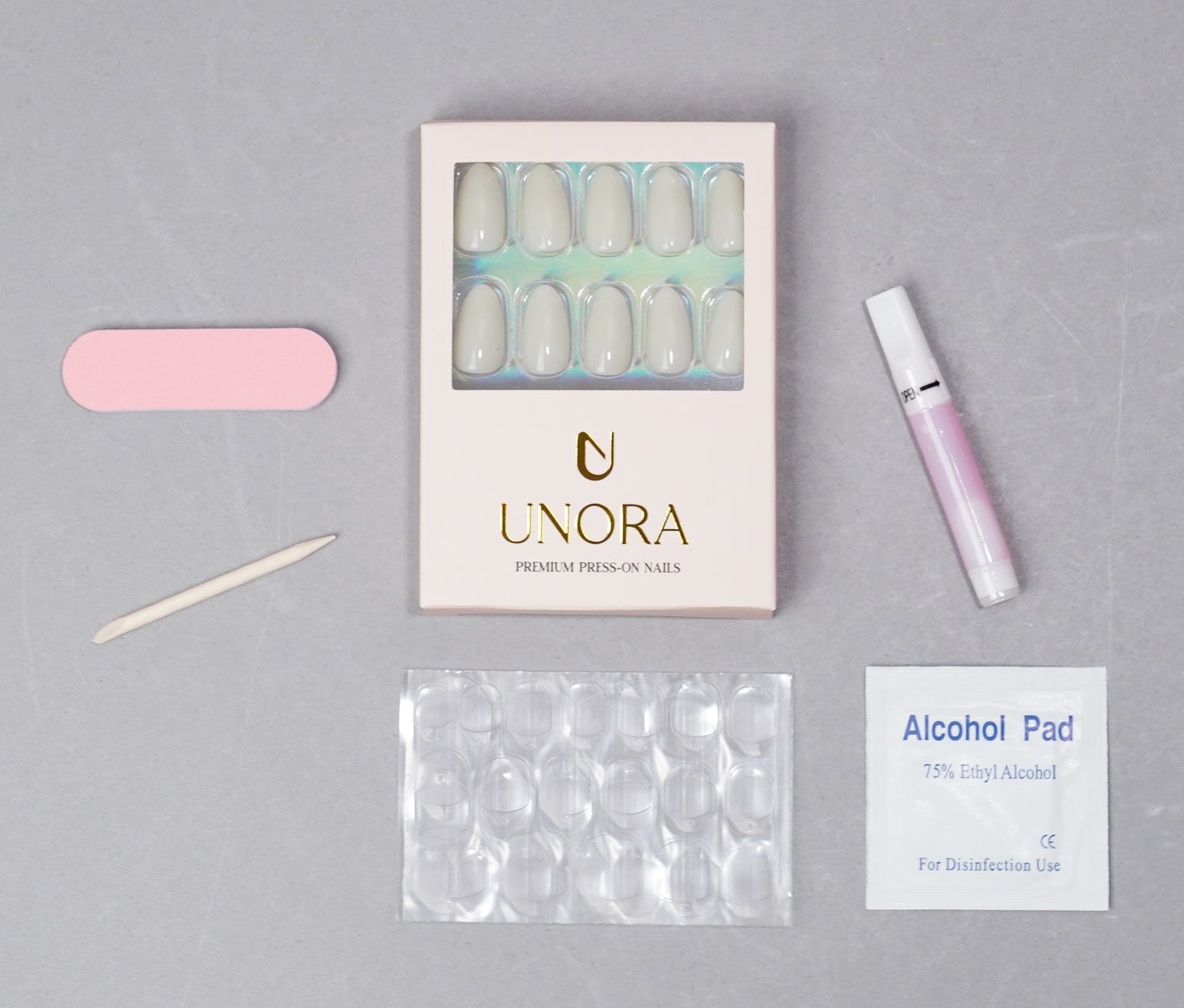 UNORA press-on nail kit laid out on a light gray background, including a box of white press-on nails, a pink nail file, a wooden nail tool, a pink gel polish tube, a packet of clear nail tips, and an alcohol pad.