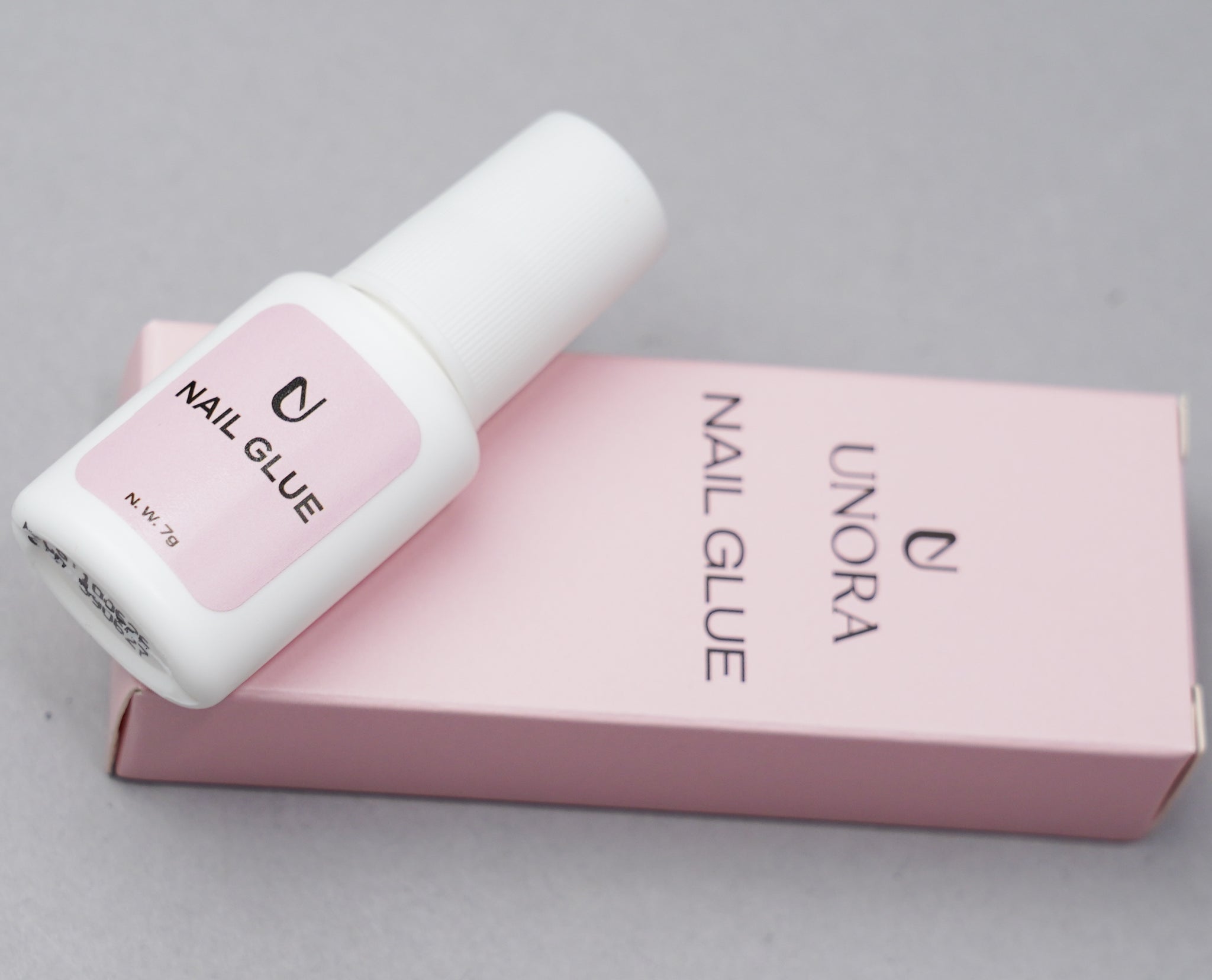 A bottle of UNORA nail glue with a white cap and a light pink label, resting diagonally on top of its matching light pink packaging box against a neutral gray background.